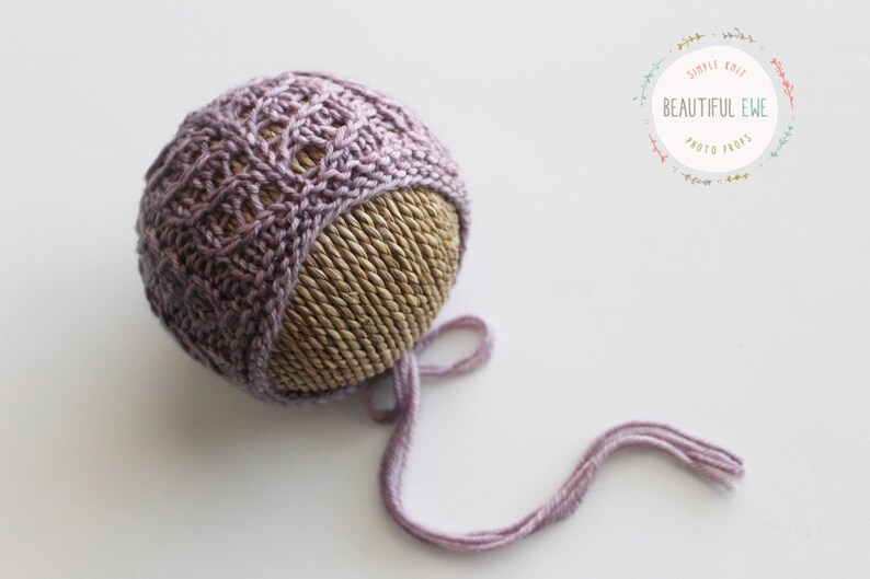 May include: A purple knitted baby bonnet with a lace pattern and a long tie. The bonnet is displayed on a woven straw ball.