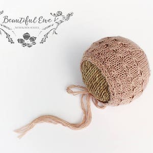 以下が含まれることがあります： ピンクのニットのベビーボンネット。編まれた麦わら帽子のつばと長いリボンが付いています。ボンネットは繊細なステッチパターンで作られており、柔らかく心地よい感触です。