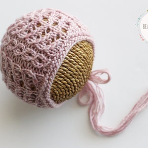 Puede incluir: Un gorro de bebé de punto rosa con una larga corbata, presentado sobre una base de paja trenzada. El gorro tiene un delicado patrón de punto de cable.