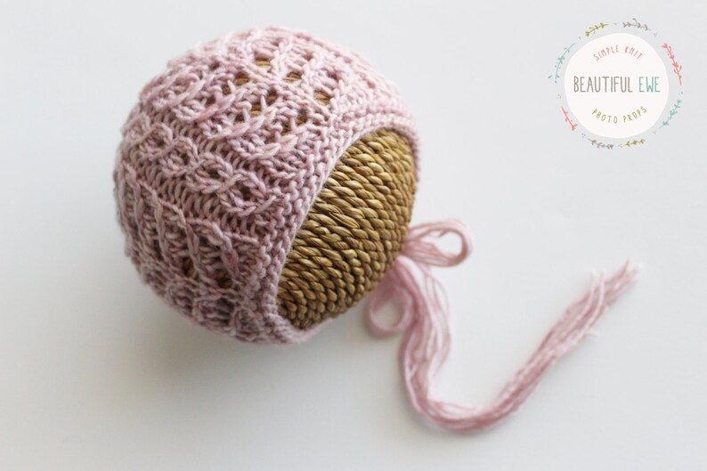 May include: A pink knitted baby hat with a long tie, displayed on a woven straw base. The hat has a delicate cable knit pattern.