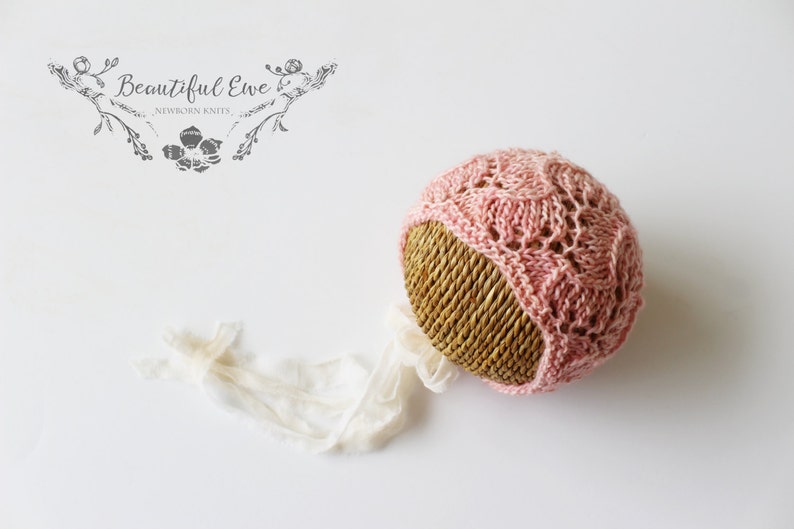 May include: A pink crocheted baby hat with a delicate lace pattern. The hat is displayed on a woven straw hat form.