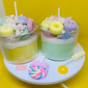 May include: Two dessert-themed candles in clear glass jars, each with a white wick. The candles have layers of yellow, green, and white wax, topped with miniature donut-shaped wax pieces in pastel colors and a crumbly topping. A white plate holds a lollipop and a yellow candy.