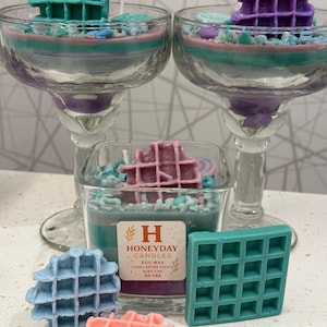 Könnte beinhalten: Mehrere Kerzen in Margaritagläsern und quadratischen Gläsern, gekrönt mit waffelförmigem Wachs. Die Kerzen haben Schichten aus türkisem, rosa und lila Wachs. Zusätzliche waffelförmige Wachsstücke in Blau, Koralle und Türkis sind ebenfalls vorhanden.