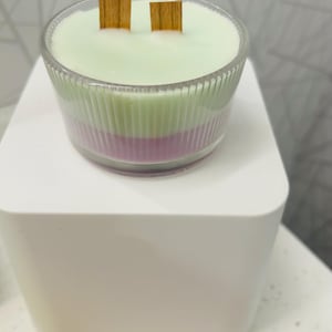 May include: A layered candle in a clear, ribbed glass container. The candle has three layers: a light green top layer, a green middle layer, and a purple bottom layer. Two wooden wicks are visible. The candle sits on a white, square pedestal.