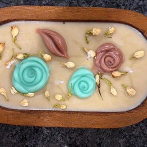 May include: A decorative candle in a wooden tray. The candle has a light beige wax base with teal and brown floral accents. Dried flower buds are scattered around the edges, adding a natural touch to the design.