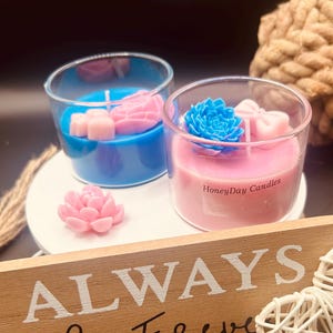 Op de afbeelding: Twee glazen kaarsen, één blauw en één roze, elk met een decoratief bloemmotief. De roze kaars heeft de tekst "HoneyDay Candles". Een houten bordje hieronder luidt "ALWAYS & Forever".