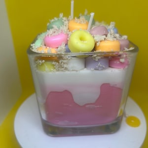 Puede incluir: Una vela decorativa con forma de postre, presentada en un vaso transparente. Presenta capas de cera rosa y blanca, adornada con trozos de cera coloridos en forma de anillo y una cobertura desmenuzada. Se ven dos mechas blancas.