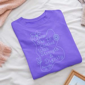 May include: A lavender crewneck sweatshirt with the text "She Is Clothed With Strength & Dignity" and Proverbs 31:25. The design includes floral and butterfly illustrations. The sweatshirt is folded on a white surface.