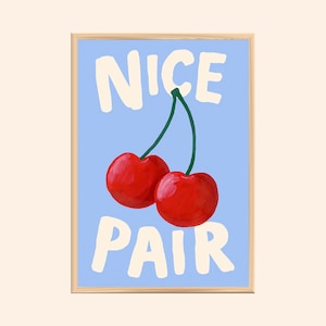 May include: A framed art print showing two red cherries with a green stem against a light blue backdrop. The words "NICE PAIR" are written in white, bold letters above and below the cherries. The frame is a light brown colour.