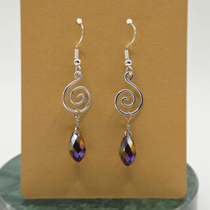 Può includere: Un paio di orecchini pendenti color argento. Ogni orecchino presenta un design a spirale e un cristallo sfaccettato a forma di goccia con sfumature iridescenti di viola, oro e marrone. Gli orecchini sono esposti su uno sfondo marrone.