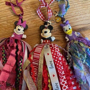 Peut inclure: Trois ornements Disney faits &agrave; la main. Les figurines de Minnie Mouse, Mickey Mouse et Daisy Duck sont attach&eacute;es &agrave; des pompons en ruban color&eacute;s. L'ornement Mickey Mouse a un ruban avec le mot "love". Les ornements sont sur une surface en bois.