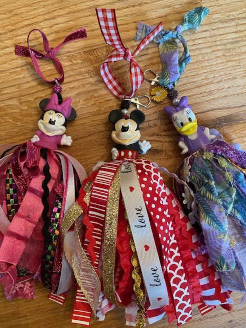 Peut inclure: Trois ornements Disney faits &agrave; la main. Les figurines de Minnie Mouse, Mickey Mouse et Daisy Duck sont attach&eacute;es &agrave; des pompons en ruban color&eacute;s. L'ornement Mickey Mouse a un ruban avec le mot "love". Les ornements sont sur une surface en bois.