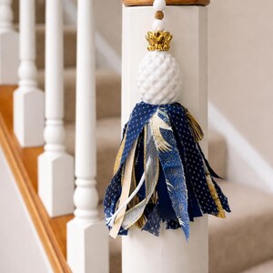 May include: A decorative tassel featuring a white pineapple-shaped top, gold detailing, and a navy and gold fabric skirt. The tassel is suspended from a wooden handrail, adding a touch of elegance to the staircase.