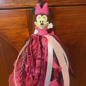 Peut inclure: Un ornement de gland sur le thème de Minnie Mouse. L'ornement présente une figurine de Minnie Mouse dans une robe et un nœud roses, avec une cascade de rubans roses, blancs et noirs. Une petite boule perlée rose est suspendue à un cordon rouge.