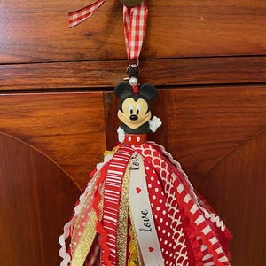 Peut inclure: Un ornement de gland Mickey Mouse avec des rubans rouges, blancs et dor&eacute;s. Le gland comprend une figurine de Mickey Mouse, une perle dor&eacute;e et un ruban vichy rouge et blanc pour l'accrocher. Le ruban blanc porte le mot "love".