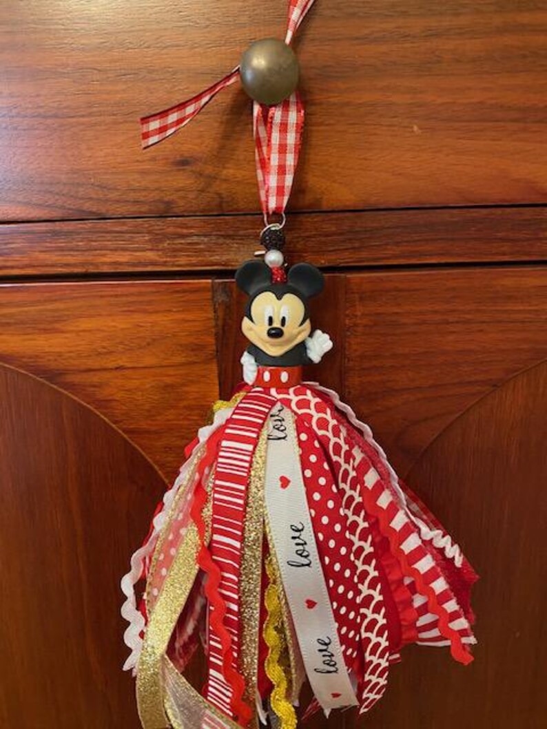 Peut inclure: Un ornement de gland Mickey Mouse avec des rubans rouges, blancs et dor&eacute;s. Le gland comprend une figurine de Mickey Mouse, une perle dor&eacute;e et un ruban vichy rouge et blanc pour l'accrocher. Le ruban blanc porte le mot "love".