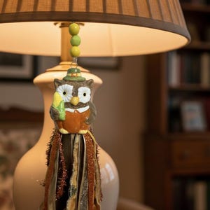 May include: A ceramic owl ornament hanging from a lamp, featuring a brown owl wearing a green hat and holding a yellow corn cob. The ornament is suspended by a string of three yellow beads and includes a decorative tassel of brown and orange ribbons.