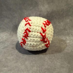 Puede incluir: Una pelota de béisbol de ganchillo, blanca con costuras rojas. La pelota es redonda y parece hecha a mano, con hilo rojo formando el patrón clásico de costuras de béisbol. Probablemente sea un objeto decorativo o un juguete.