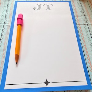 Puede incluir: Un bloc de notas rectangular con un borde azul brillante, que presenta las letras JT impresas en negro en la parte superior y un gráfico de estrella de cuatro puntas en la parte inferior. Un lápiz de madera amarillo con goma rosa está colocado verticalmente en el lado izquierdo.