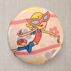 May include: A round button featuring a cartoon character with blonde hair and pink glasses. The character is wearing a red top and blue pants, with a background of pink stripes and flowers. The button has a glossy finish.