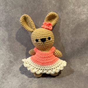 May include: A handmade crochet bunny doll with a light brown body, long ears, and a small pink flower on its head. The bunny wears a pink and white dress. The doll has black eyes and a black nose. The background is a solid gray color.