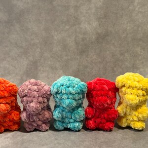 Puede incluir: Cinco osos de ganchillo hechos a mano en fila. Los osos son de varios colores: naranja, morado, turquesa, rojo y amarillo. Los osos tienen una apariencia texturizada y afelpada y están de pie.