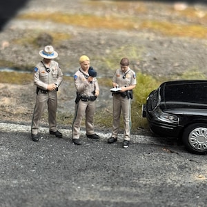 May include: Three miniature police officer figurines standing on a textured gray surface next to the front portion of a black car. The figures wear tan uniforms with dark belts and accessories. One officer holds a radar gun, another holds a notepad.