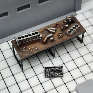 May include: A miniature workshop scene with a brown workbench and various engine parts. The workbench has a metal frame and is covered with small tools and engine components. A small metal plaque with engine details sits on the floor. The scene is set against a gray backdrop.