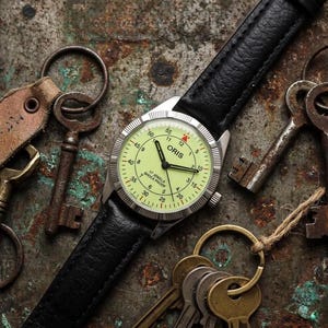 Op de afbeelding: Een vintage horloge met een lichtgroene wijzerplaat en een zwarte leren band. Het horloge wordt omringd door antieke sleutels en sleutelhangers op een getextureerd, roestig metalen oppervlak. De wijzerplaat toont het merk "ORIS".