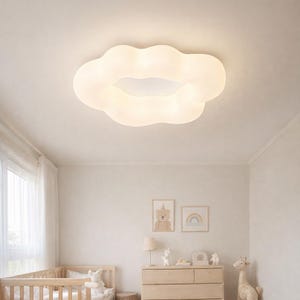 May include: A cloud-shaped ceiling light in a nursery. The light fitting is a soft white colour and is mounted on a white ceiling. The room includes a cot, chest of drawers, and artwork, creating a calming atmosphere.