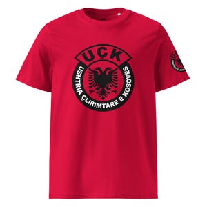 Może przedstawiać: Czerwona koszulka z czarnym emblematem przedstawiającym dwugłowego orła i napisem "UÇK Ushtria Çlirimtare e Kosovës". Ten sam emblemat powtarza się na rękawie. Koszulka wykonana jest z miękkiego materiału.