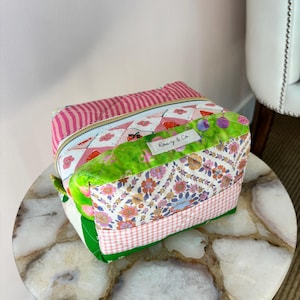 May include: A set of quilted cosmetic bags in various patterns. The top bag has pink and white stripes, while the bottom bag features floral and gingham patterns. The bags are sitting on a decorative table.