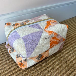 Peut inclure: Une pochette matelassée à fermeture éclair présentant un motif patchwork de panneaux de tissu triangulaires dans des tons orange, violet et crème avec divers motifs. Une petite étiquette en tissu avec le texte Rosy & Co. est cousue sur le côté.