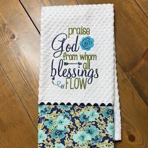 May include: White kitchen towel with a blue floral border. The towel has an embroidered message that reads "Praise God from whom all blessings flow" with a blue flower and arrow.