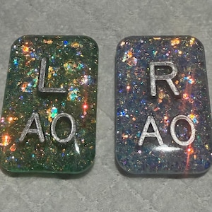 May include: Two rectangular resin pieces with glitter and silver lettering. One is green with the letters "L AO", the other is blue with "R AO". The pieces are likely for a craft or jewelry project.