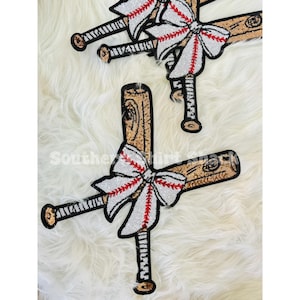 Puede incluir: Aplique de bate de béisbol con un lazo de lentejuelas blanco y rojo. Los bates son marrones con detalles dorados y mangos negros. El texto "Southern Shirt Shack" es visible en la imagen.