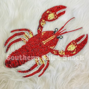 May include: A red beaded lobster design with yellow accents and the text "Southern Shirt Shack" on a white fluffy background. The lobster has detailed claws, antennae, and a segmented body.