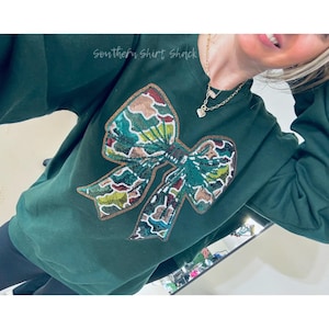 May include: Dark green sweatshirt with a large sequined bow design in shades of green, brown, and gold. The text "Southern Shirt Shack" is printed in cursive on the top left. The sweatshirt is a casual top.