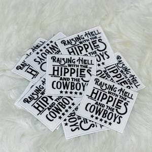 May include: A collection of square white fabric patches featuring black embroidered text that reads Raising Hell with the Hippies and the Cowboys, accented with a row of five small black stars at the bottom of each individual patch design.