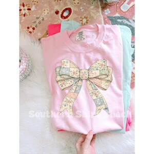 May include: A light pink Comfort Colors sweatshirt featuring a large bow design with a patchwork pattern of floral and pastel squares. The bow is the focal point, with the text "Southern Star Shack" visible at the bottom.