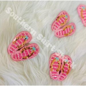 May include: Pink sequined flip-flops with gold and blue details. The flip-flops feature a striped pattern and a star on the toe. The text "Southern Shirt Shack" is in the background.
