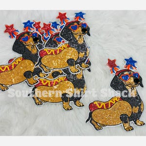 May include: A collection of sequined patches featuring dachshund dogs dressed as hot dogs. Each dog wears heart-shaped sunglasses and has red and blue star-shaped accents above its head. The patches are black, gold, red, and blue with glitter.