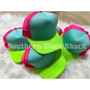 May include: A stack of trucker hats with a neon green brim, a teal crown, and a pink mesh back. The hats have the text "Southern Shirt Shack" printed on the front.