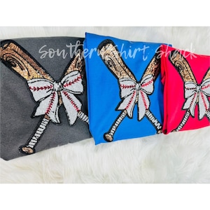 May include: Three baseball-themed shirts in gray, blue, and red. Each shirt features a sequined baseball bat design with a bow and baseball stitching. The text "Southern Shirt Shack" is visible on the gray shirt.