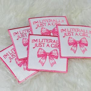 May include: A collection of square white fabric patches featuring pink embroidered text that reads I'M LITERALLY JUST A GIRL above a pink embroidered bow graphic. The patches have a pink stitched border and are arranged in a overlapping cluster.