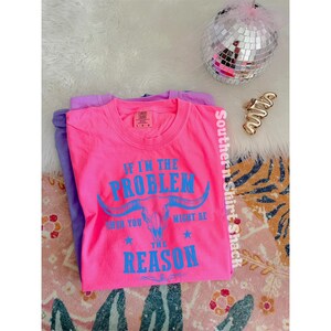 May include: A bright neon pink t-shirt featuring a blue graphic of a longhorn skull and the text IF I'M THE PROBLEM THEN YOU MIGHT BE THE REASON. The shirt has a crew neckline and a Comfort Colors brand label on the inside of the collar.