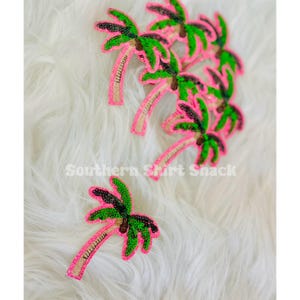 May include: Embroidered palm tree patches in pink, green, and tan. The patches are embellished with sequins and beads. The text "Southern Shirt Shack" is printed in white. The patches are arranged on a white, fluffy surface.