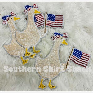 May include: Three sequined goose patches in beige with yellow beaks and feet. Each goose wears a blue, white, and red star-patterned bow and holds a sequined American flag. The text Southern Shirt Shack is overlaid in white at the bottom.