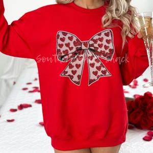 May include: A red sweatshirt featuring a large bow design. The bow is embellished with red heart-shaped sequins and a white outline. The text "Southern Shack" is visible on the sweatshirt. The overall theme is Valentine's Day.