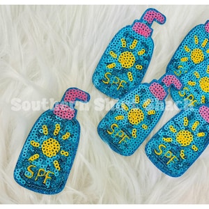 May include: A collection of blue sequined sunscreen bottle patches. Each patch features a yellow sun design and the letters "SPF". The bottle tops are pink.